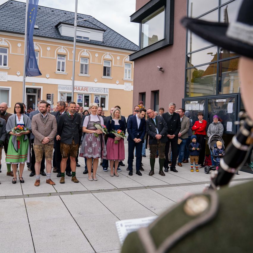 Eine Gruppe von Menschen steht auf einem gepflasterten Bereich vor einem Gebäude. Sie tragen alle traditionelle Kleidung und einige halten Blumen. Ein Mann mit Hut und Musikinstrument steht vor ihnen.