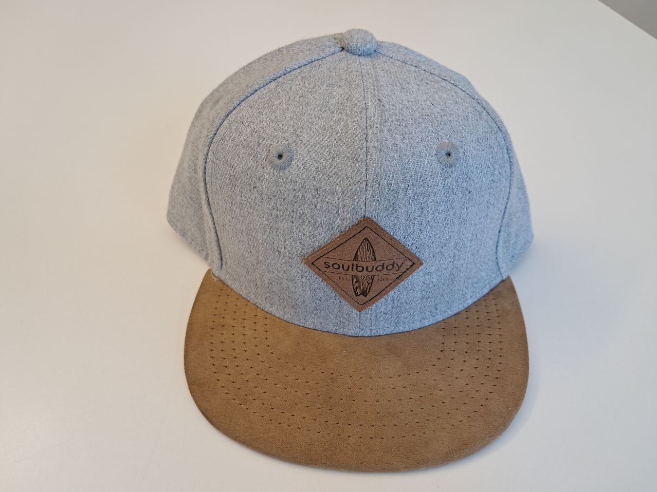 Eine graue und braune Baseballkappe mit einem braunen Logo-Patch auf der Vorderseite, der ein Surfbrett und den Text 'soulbuddy' zeigt. Die Kappe hat eine flache Krempe und ist von vorne gezeigt.