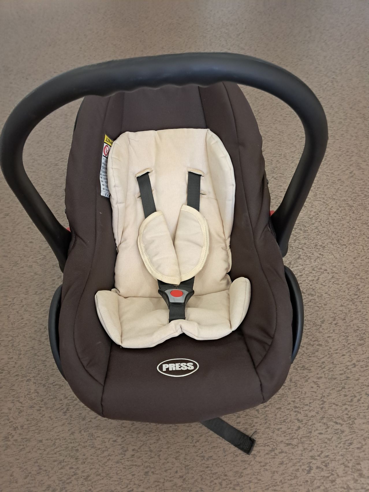 A brown and beige infant car seat with a black handle is shown. It has a beige padded seat, a black seat belt, and the word PRESS in white on the front.