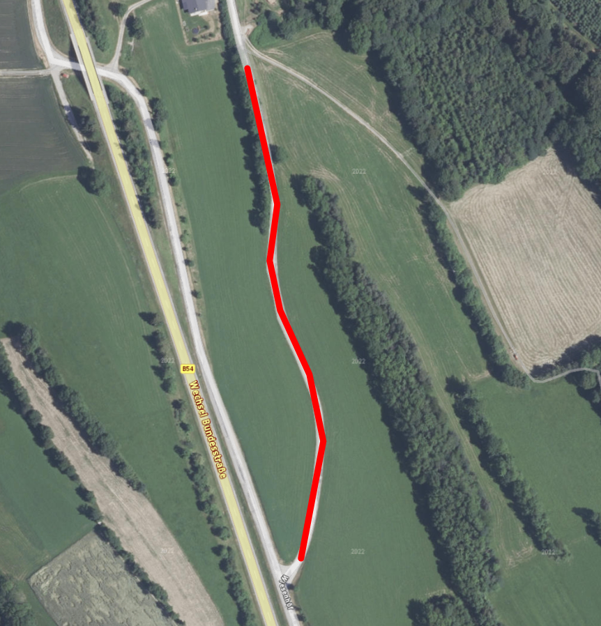 Eine Luftaufnahme eines ländlichen Gebiets mit einer roten Linie, die einen Weg durch Felder und Bäume markiert. Der Weg führt zu einer Straße mit der Bezeichnung B54.