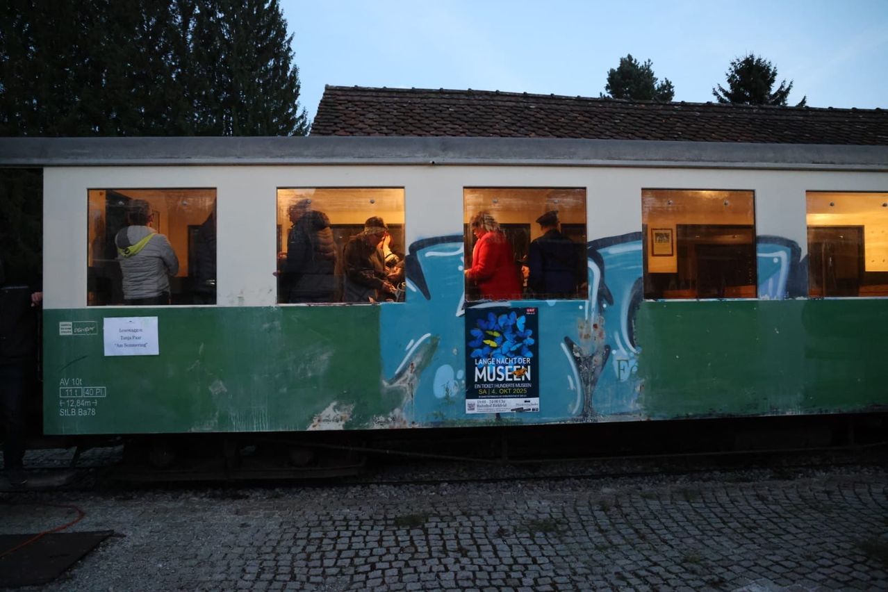 Ein weiß-grüner Zug mit Graffiti, der ein Plakat mit Lange Nacht der Museen zeigt. Menschen sind im Inneren des Zuges zu sehen.