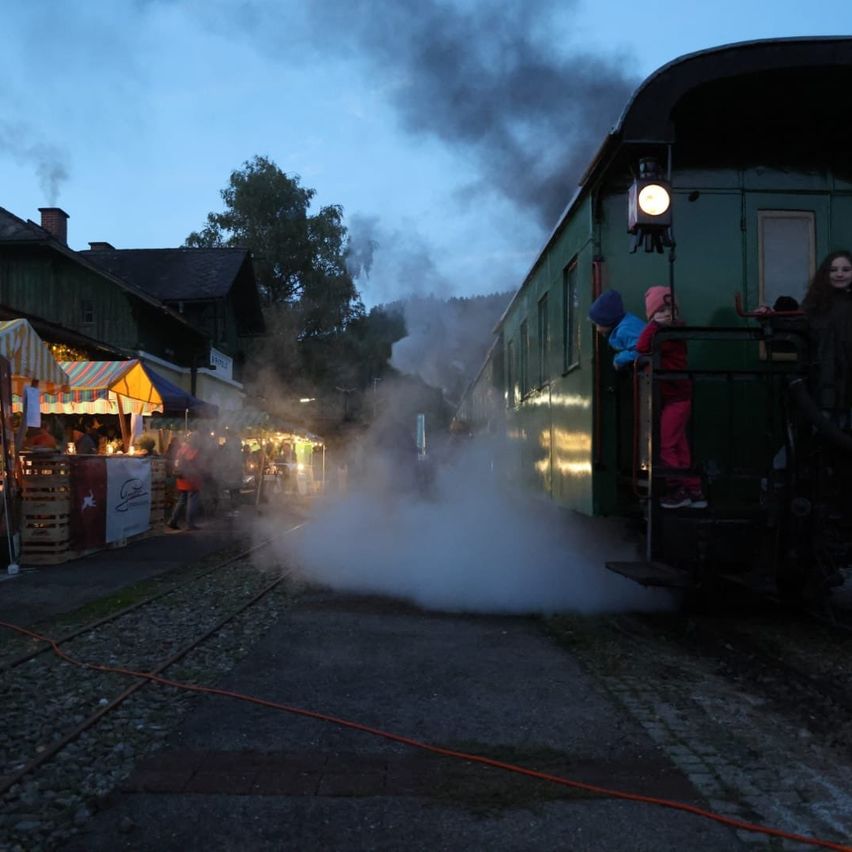Eine Dampflokomotive emittiert Dampf und Rauch, während sie auf einer Bahnstrecke fährt, wobei Kinder aus dem Fenster schauen. In der Nähe versammeln sich Menschen um ein farbenfrohes Zelt mit Tischen.