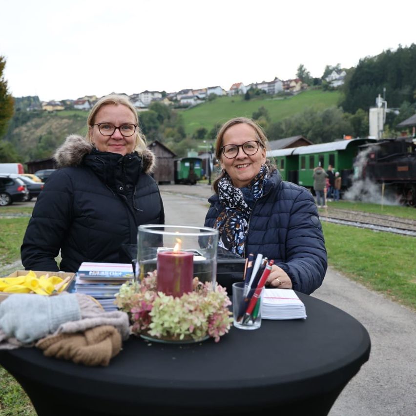 Zwei Frauen in Winterkleidung stehen bei einem Tisch mit Gegenständen und einer Kerze, in der Nähe einer auf den Gleisen fahrenden Lokomotive, mit einer Stadt im Hintergrund.