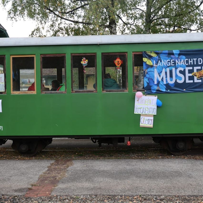 Ein grüner Vintage-Zugwagen steht in einem Außenbereich geparkt, mit Bannern und Luftballons daran befestigt. Der Zug hat Glasfenster und einige Menschen sind im Inneren. Im Hintergrund sind Bäume zu sehen.