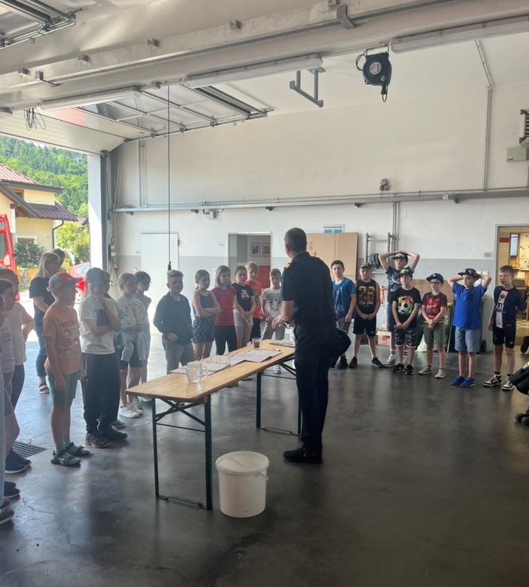 Ein Feuerwehrmann steht mit einer Gruppe von Kindern in einer Garage an einem Tisch. Papiere und Gläser liegen auf dem Tisch.