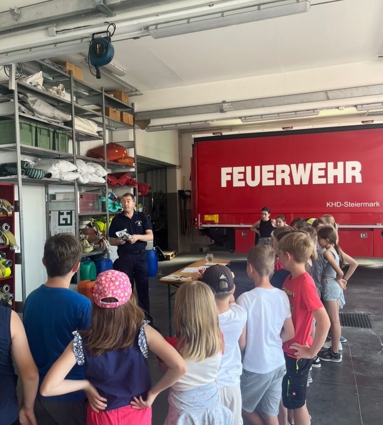Ein Feuerwehrmann hält eine Präsentation für eine Gruppe von Kindern in einer Feuerwache. Die Kinder stehen in einer Reihe und hören dem Feuerwehrmann zu.