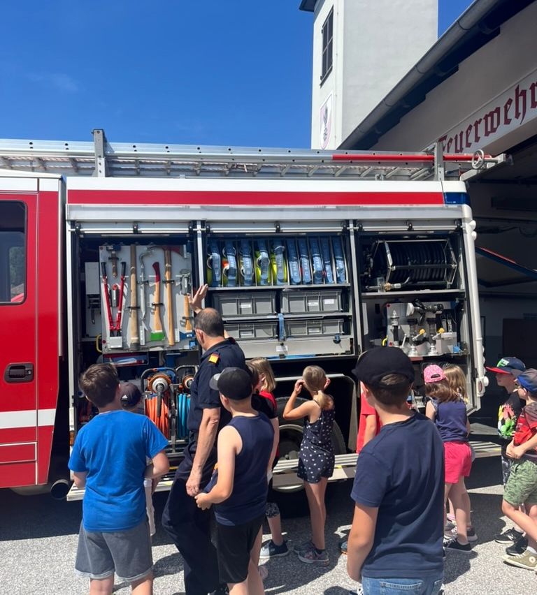 Ein Feuerwehrmann zeigt Kindern die Ausrüstung in einem Feuerwehrauto. Sie stehen vor dem Fahrzeug, einige schauen hinein, während andere zuschauen.