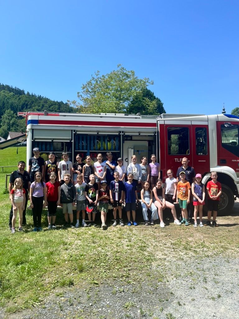 Eine Gruppe von Kindern und Erwachsenen posiert für ein Foto vor einem Feuerwehrauto an einem sonnigen Tag.