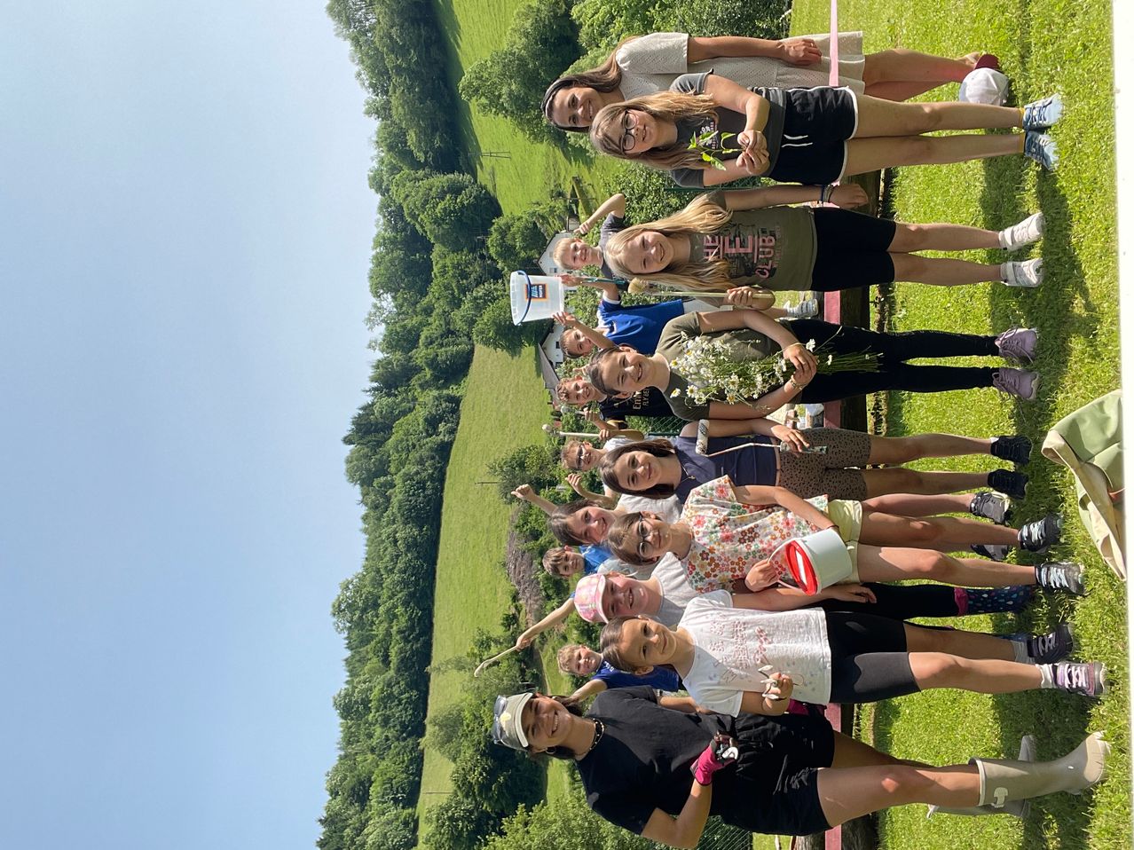 Eine Gruppe junger Mädchen und Jungen steht auf einem grünen Feld. Sie lächeln und scheinen für ein Foto zu posieren. Einige von ihnen halten Blumen und Eimer. Der Hintergrund zeigt ein grünes Feld mit Bäumen.