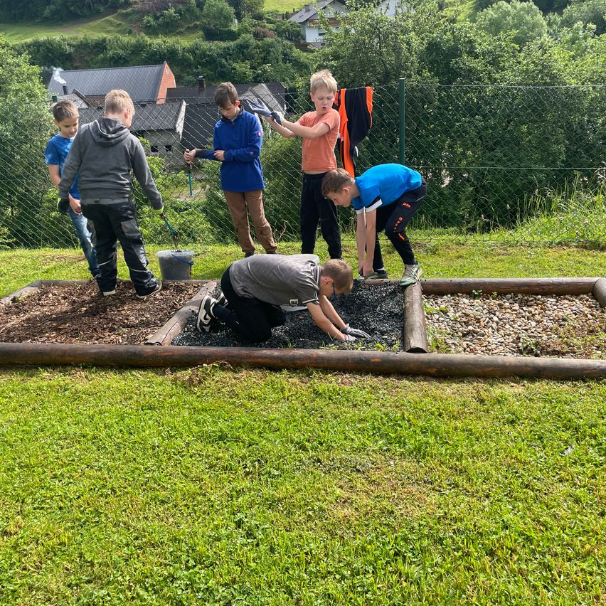 Eine Gruppe von Jungen pflanzt ein Gartenbeet in einem Hof. Sie tragen Handschuhe und arbeiten mit Werkzeug. Im Hintergrund befinden sich Häuser und Bäume.