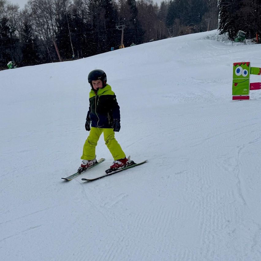 Ein Kind  skiing auf einer verschneiten Piste mit einem gelben und schwarzen Jacke, einem schwarzen Helm, Handschuhen und gelben Skihosen. Ein grüner Maskottchen steht auf der rechten Seite der Piste.