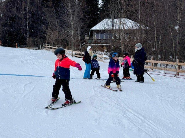 Eine Gruppe von Kindern und Erwachsenen skifahrern auf einer verschneiten Piste mit einem Haus im Hintergrund.