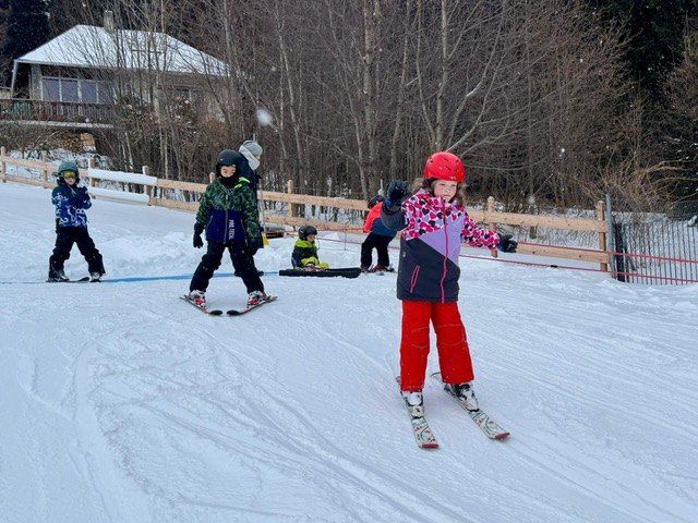 Mehrere Kinder üben das Skifahren auf einer verschneiten Piste. Ein Kind trägt einen roten Helm und rote Hosen. Dahinter stehen Bäume und ein Haus.
