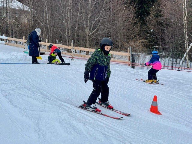Eine Gruppe von Menschen  Skifahren den Schneehang hinunter. Eine Person benutzt einen Snowboard. Bäume und ein Zaun umgeben das Gebiet.