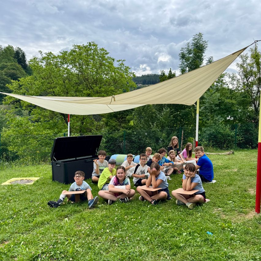 A group of children sitting under a large beige canopy on a grassy field, some wearing glasses, with a black storage box nearby.