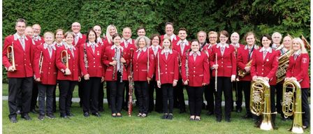 Eine Gruppe von Musikern in roten Uniformen, die für ein Foto auf einem Rasenfeld mit Bäumen im Hintergrund posieren. Sie halten verschiedene Musikinstrumente.