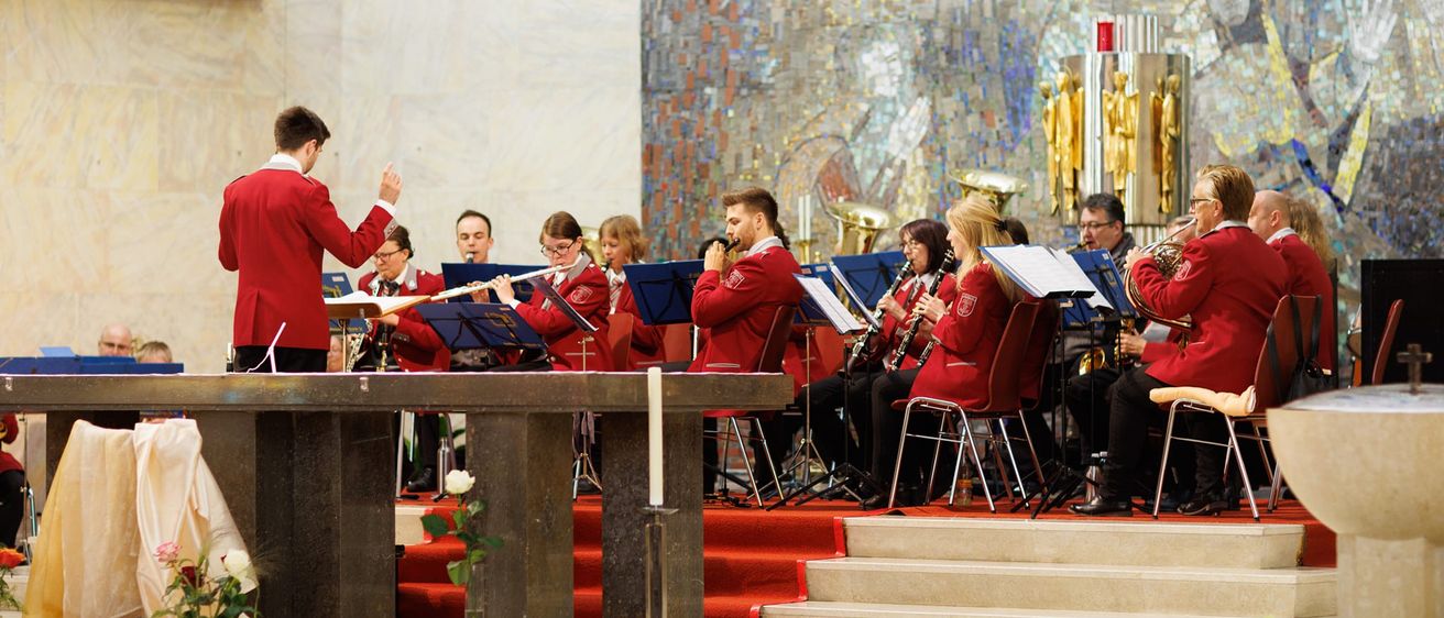 Ein Blasorchester in roten Uniformen spielt in einer Kirche, mit einem Dirigenten vorne, umgeben von verschiedenen Musikinstrumenten.
