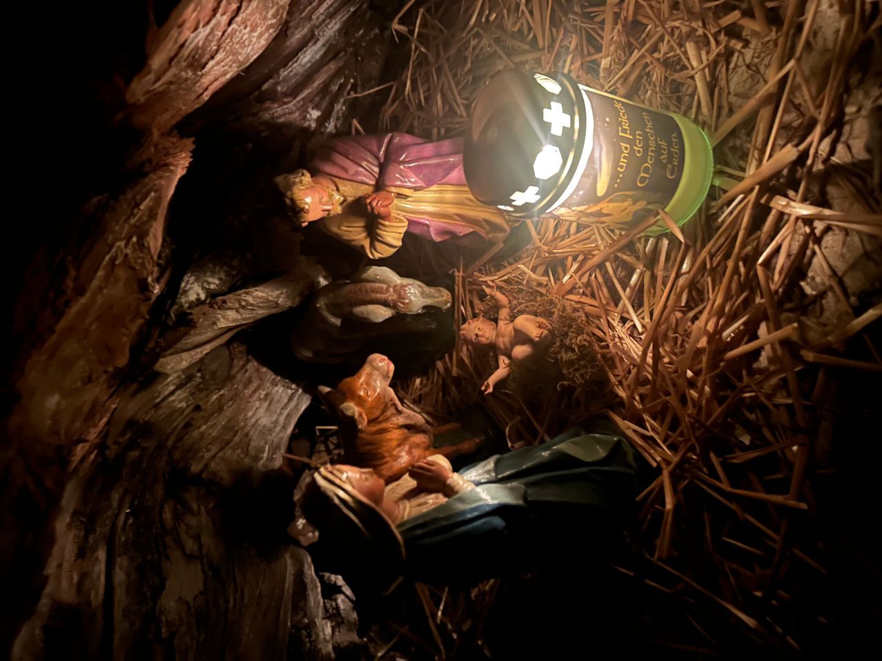 A nativity scene with figurines of Mary, Joseph, and baby Jesus in a manger, illuminated by a candle. A small green bottle is placed beside the manger.