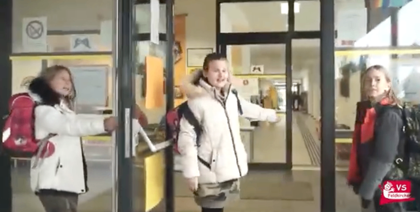 Drei Mädchen in Wintermänteln stehen in einem Schulkorridor, eines hält einen Türgriff, ein anderes lächelt und das dritte schaut voraus.
