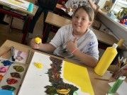 Ein junges Mädchen sitzt an einem Tisch und malt ein Bild mit gelber und grüner Farbe. Es gibt mehrere Farbflaschen und Pinsel auf dem Tisch. Der Raum hat Holztische und Stühle.