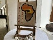 Ein Staffelei mit einem Gemälde Afrikas in warmen Farben steht auf einem Tisch. Das Gemälde zeigt das Wort 'Hakuna Mata' in fetten Buchstaben.