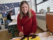 Ein junges Mädchen in einem roten Sweatshirt malt ein Bild auf einer Leinwand in einem Kunstraum, während eine andere Frau im Hintergrund arbeitet.