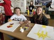 Zwei junge Mädchen sitzen an einem Tisch mit Leinwänden und malen mit verschiedenen Farben. Andere Schüler arbeiten im Hintergrund. Der Raum ist gut beleuchtet, und die Wände sind mit Postern dekoriert.