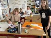 In einem Klassenzimmer sitzen ein Junge und zwei Mädchen an einem Schreibtisch. Der Junge malt auf einer großen Leinwand. Die Mädchen beobachten. Bücher und Malutensilien sind auf den Schreibtischen.