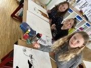 Drei Mädchen sitzen an einem Tisch und beschäftigen sich mit einer Kunstaktivität. Sie verwenden Pinsel und Farbe auf großen Papierbögen, wobei eines der Mädchen ein graues Langarmshirt trägt.