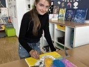 Eine junge Frau in einem schwarzen Shirt und Jeans malt ein lebhaftes Bild auf einer Leinwand in einem Raum mit Holzböden und Bücherregalen.