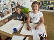 Zwei Mädchen sitzen an einem Tisch, jedes mit einem großen Blatt Papier. Ein Mädchen zeichnet mit einem Stift, das andere hält eine Schere. Verschiedene Malutensilien liegen auf dem Tisch.