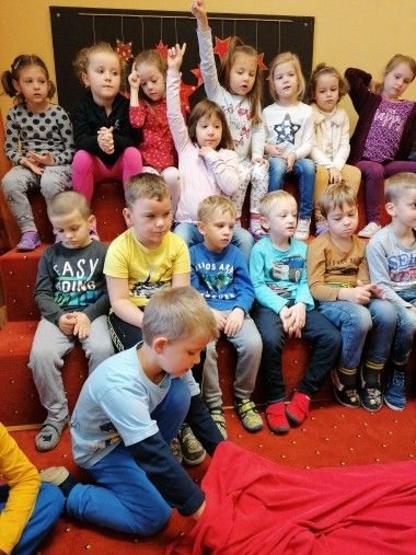 Eine Gruppe von Kindern sitzt auf rotem Teppich, einige auf Treppenstufen, mit überraschten Gesichtern. Ein Junge kniet mit einem roten Tuch.