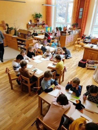 Kinder sitzen an Tischen in einem Klassenzimmer und beschäftigen sich mit Zeichenaktivitäten. Das Klassenzimmer hat Holzböden und Wände. Einige Erwachsene sind anwesend.