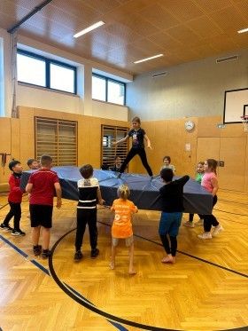 Eine Gruppe von Kindern spielt auf einem Trampolin in einer Turnhalle mit Holzböden und einem Basketballplatz.