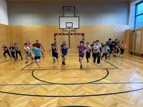 Eine Gruppe von Kindern läuft in einer Turnhalle mit einem Basketballkorb an der Wand. Sie tragen Turnschuhe und einige haben Armbänder.