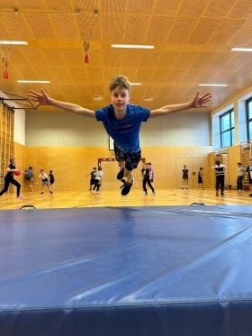Ein junger Junge in einem blauen Shirt springt auf einer blauen Matte in einer Turnhalle, mit anderen im Hintergrund.