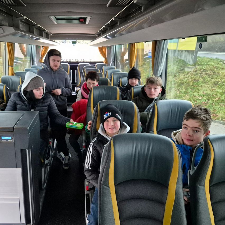 Eine Gruppe junger Jungen und ein Mann sitzen in einem Bus mit gelben und grauen Sitzen. Sie scheinen in ein Gespräch vertieft zu sein.