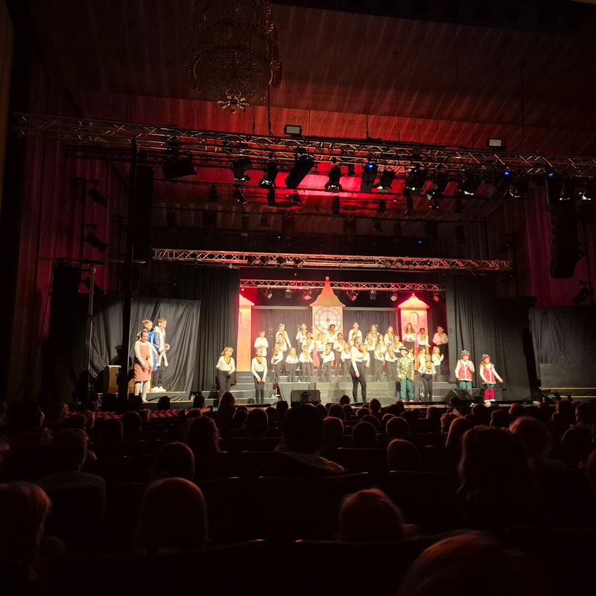 Ein Chor in passenden Kostümen tritt auf der Bühne unter roten Lichtern in einem Theater auf.
