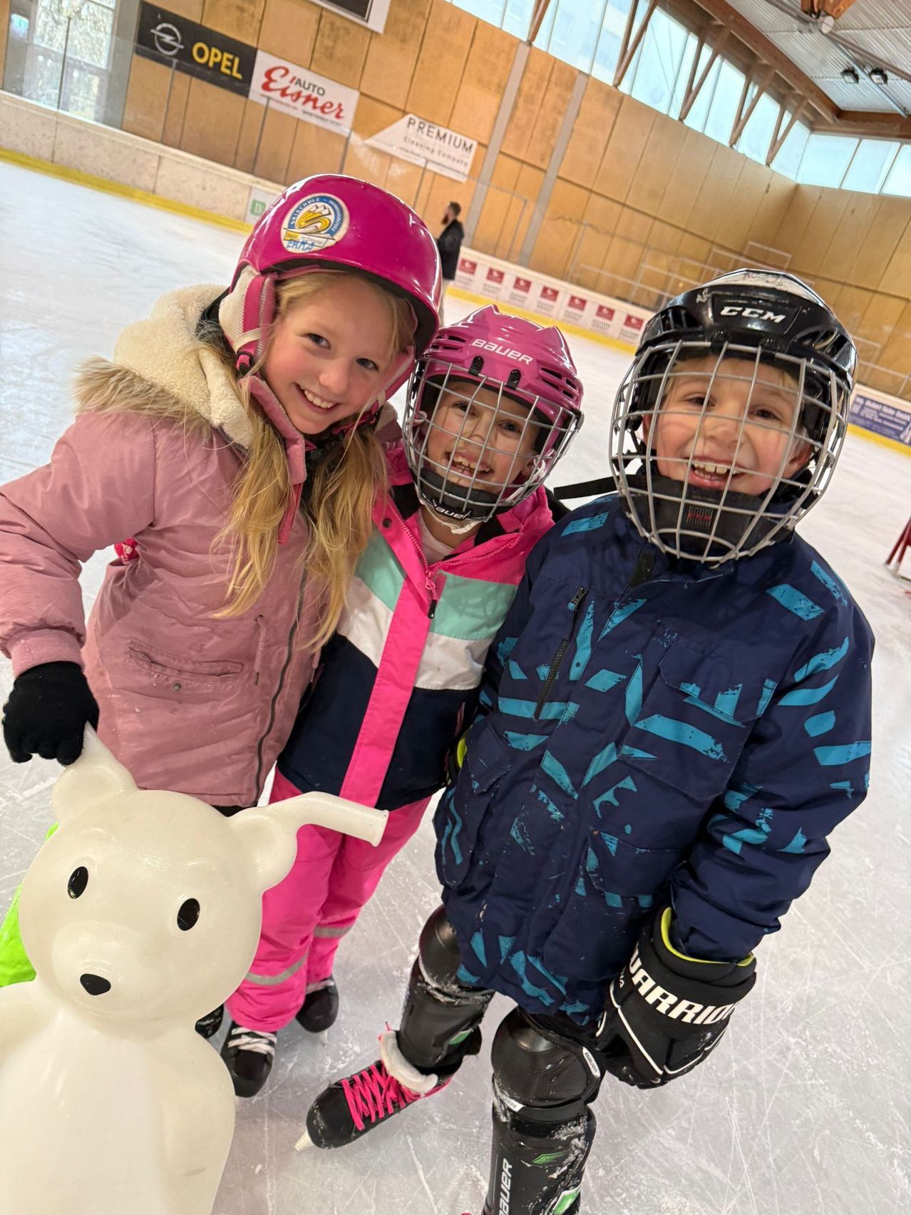 Drei Kinder, alle mit Helmen und Winterkleidung bekleidet, stehen auf einer Eisbahn. Eines hält eine weiße Bärenskulptur, ein anderes hat einen Hockeyschläger. Ein viertes Kind steht im Hintergrund.