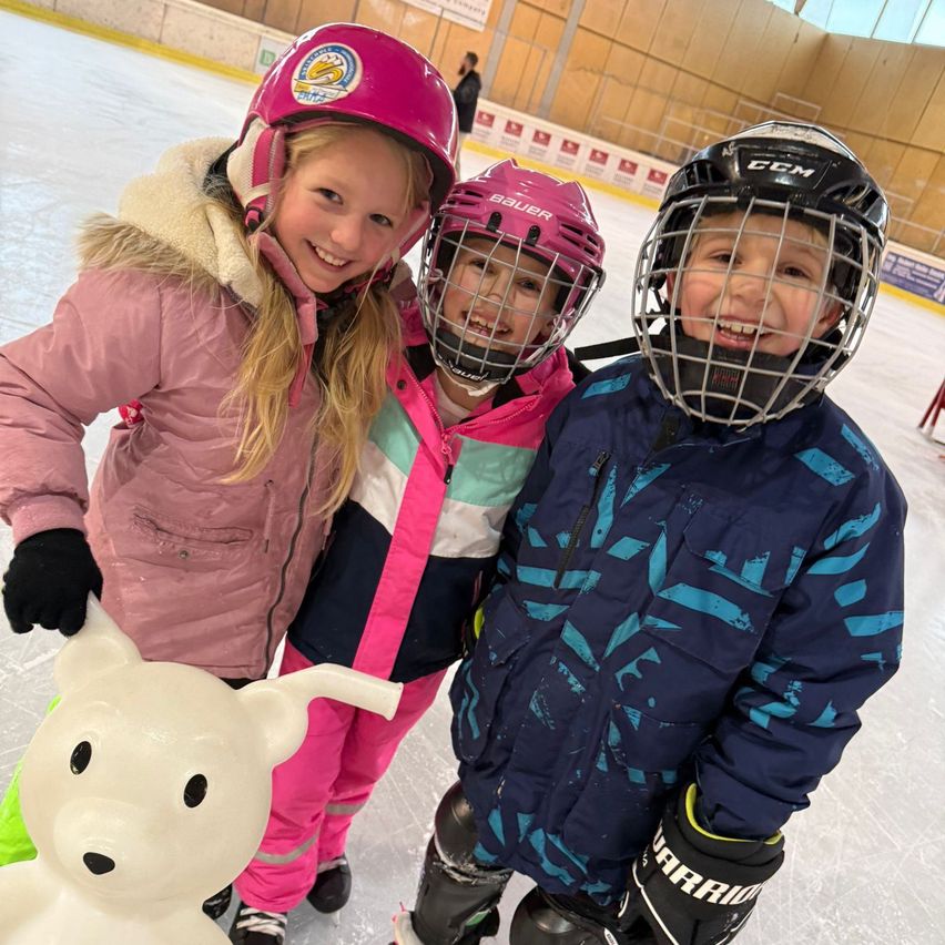 Drei Kinder, alle mit Helmen und Winterkleidung bekleidet, stehen auf einer Eisbahn. Eines hält eine weiße Bärenskulptur, ein anderes hat einen Hockeyschläger. Ein viertes Kind steht im Hintergrund.