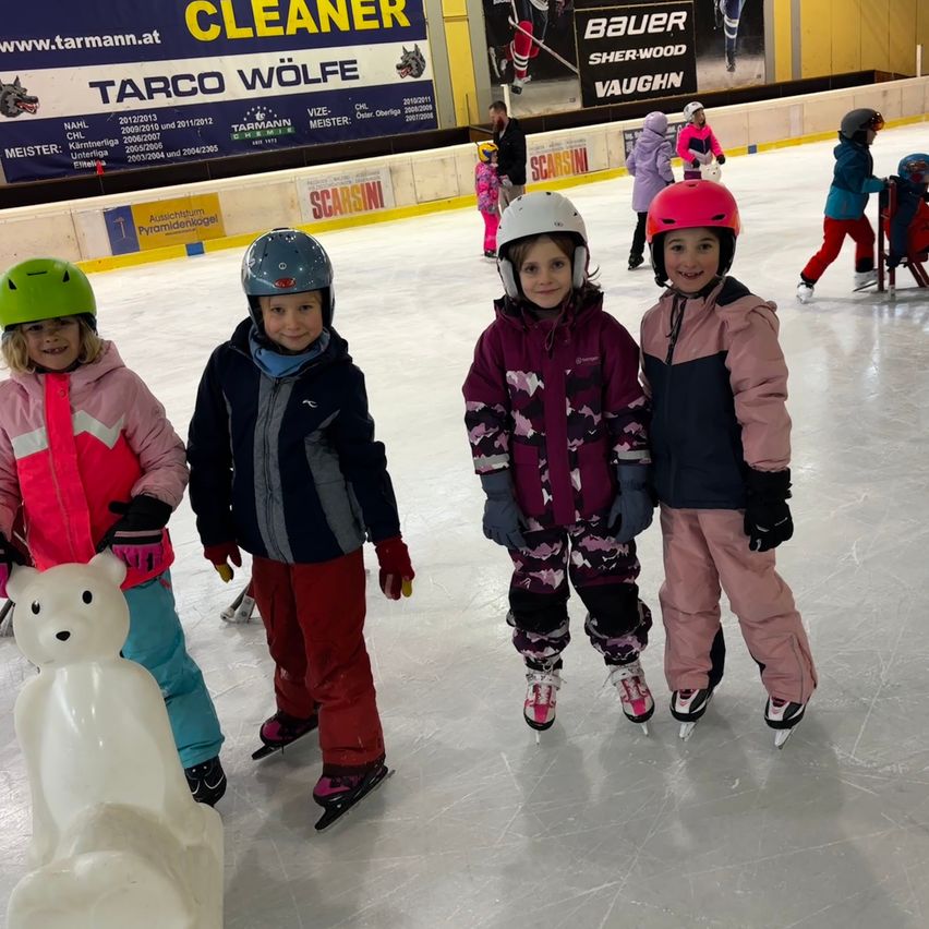 Mehrere Kinder mit Eislaufschuhen und Helmen stehen auf einer Eisbahn, lächeln und posieren für ein Foto. Eine weiße Bärenstatue ist im Vordergrund.