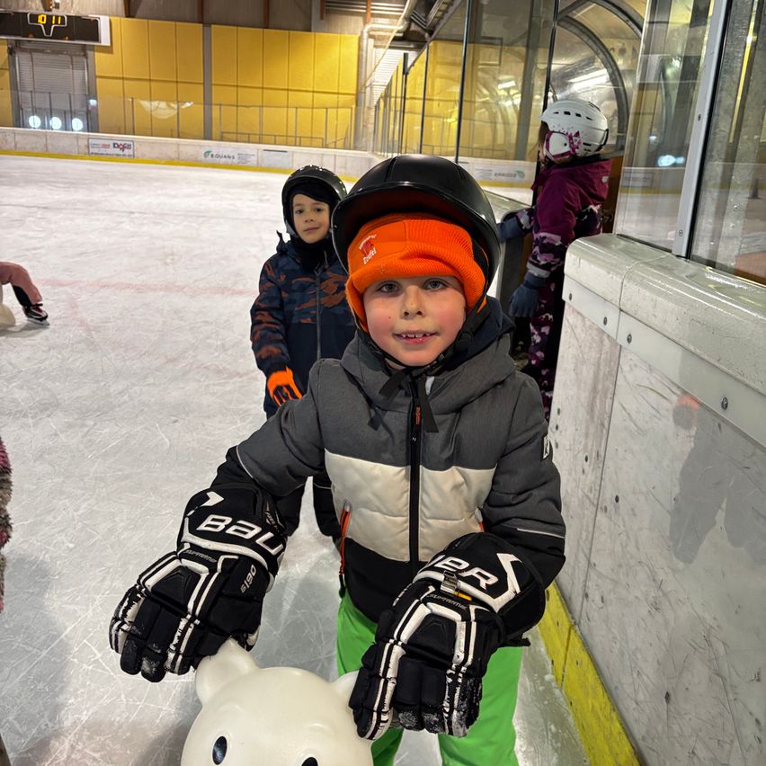 Ein junger Junge in Eishockeyausrüstung lächelt in einer Eishalle. Ein anderes Kind steht hinter ihm. Im Hintergrund findet ein Eishockeyspiel statt.