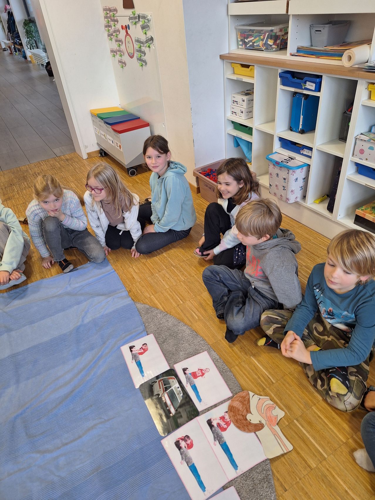 Eine Gruppe von Kindern sitzt auf dem Boden in einem Klassenzimmer und beschäftigt sich mit Lernmaterialien. Ein Kind lächelt, während ein anderes nachdenklich aussieht. Sie sind von Lagerregalen und einer ordentlichen Umgebung umgeben.