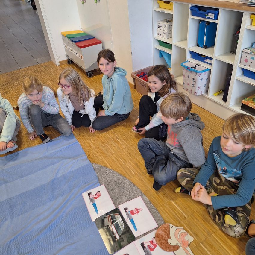 Eine Gruppe von Kindern sitzt auf dem Boden in einem Klassenzimmer und beschäftigt sich mit Lernmaterialien. Ein Kind lächelt, während ein anderes nachdenklich aussieht. Sie sind von Lagerregalen und einer ordentlichen Umgebung umgeben.