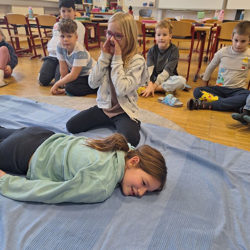 Ein Klassenzimmer mit Kindern, die in einem Kreis auf einer blauen Decke sitzen, ein Mädchen liegt, ein anderes sitzt mit Brille und ein Junge trägt Hausschuhe.