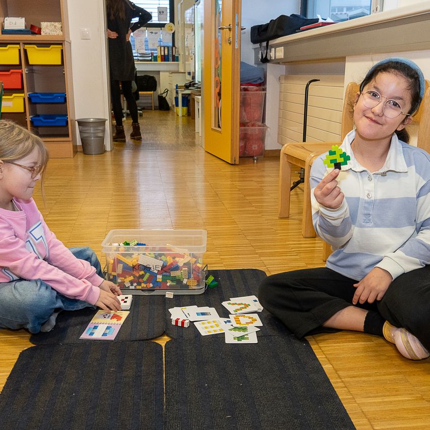 Zwei Mädchen sitzen auf dem Boden und spielen mit Puzzles. Ein Mädchen hält ein Lego-Stück in der Hand. Neben ihnen steht eine Aufbewahrungsbox voller Legos.