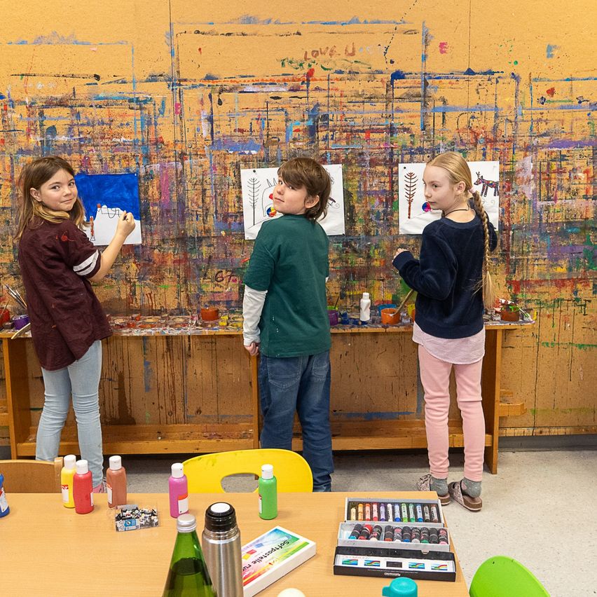 Drei Kinder malen auf einer großen Wand, die mit bunter Farbe bedeckt ist. Sie stehen vor einem Holztisch mit Farbflaschen und Pinsel. Die Wand hinter ihnen hat verschiedene Zeichnungen und Designs.