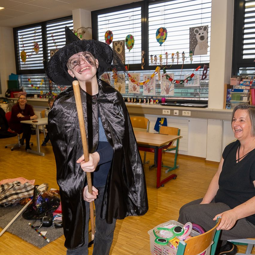Ein junger Junge, verkleidet als Hexe mit Hut und Umhang, steht mit einem Stock in einem Klassenzimmer. Eine Frau sitzt auf einem Stuhl in der Nähe und lächelt.