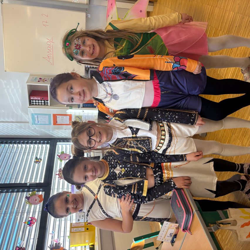 Fünf junge Mädchen in bunten Kostümen posieren für ein Foto im Klassenzimmer. Der Raum ist mit Spielzeug und Büchern dekoriert.