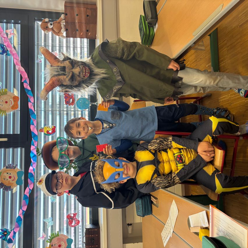 Mehrere Kinder posieren für ein Foto in einem Klassenzimmer. Eines trägt eine Maske, ein anderes hat einen Gürtel, und im Hintergrund hängen Dekorationen.